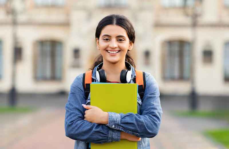 A student with a smiling face is holding a blue notebook